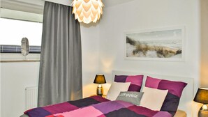 Een slaapkamer met een bed, twee bedlampen, een raam met gordijnen, een aan de muur hangend schilderij en een radiator.
