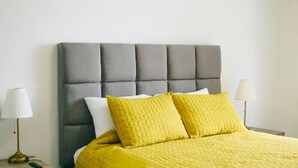 Una cama con cabecera tapizada de gris, dos almohadas blancas y dos almohadas amarillas. Hay dos mesitas de noche con lámparas a ambos lados de la cama.
