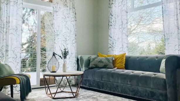 Una sala de estar con un sofá gris, cojines amarillos y verdes, una mesa de centro redonda y un jarrón con plantas.