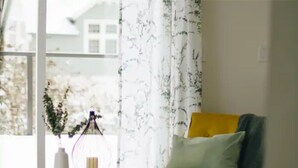 Una habitación con un sofá amarillo, una mesa redonda con un jarrón y un farol, y una ventana con cortinas estampadas.