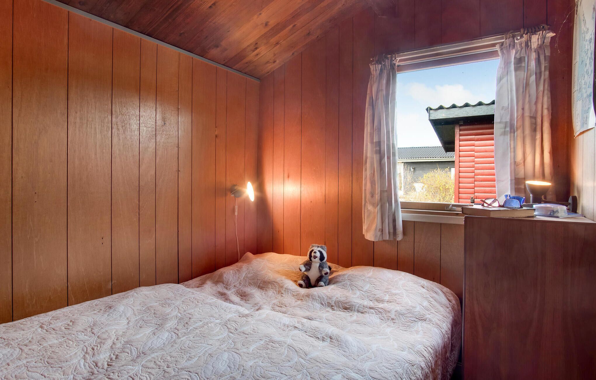 A wooden room with a bed, a window with curtains, and a desk with a lamp and some items on it.