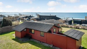 A red cabin with a sloped roof, situated on a grassy area with a view of a body of water and other buildings in the background. - 1 bedroom awesome home in Kalundborg (Kalundborg)