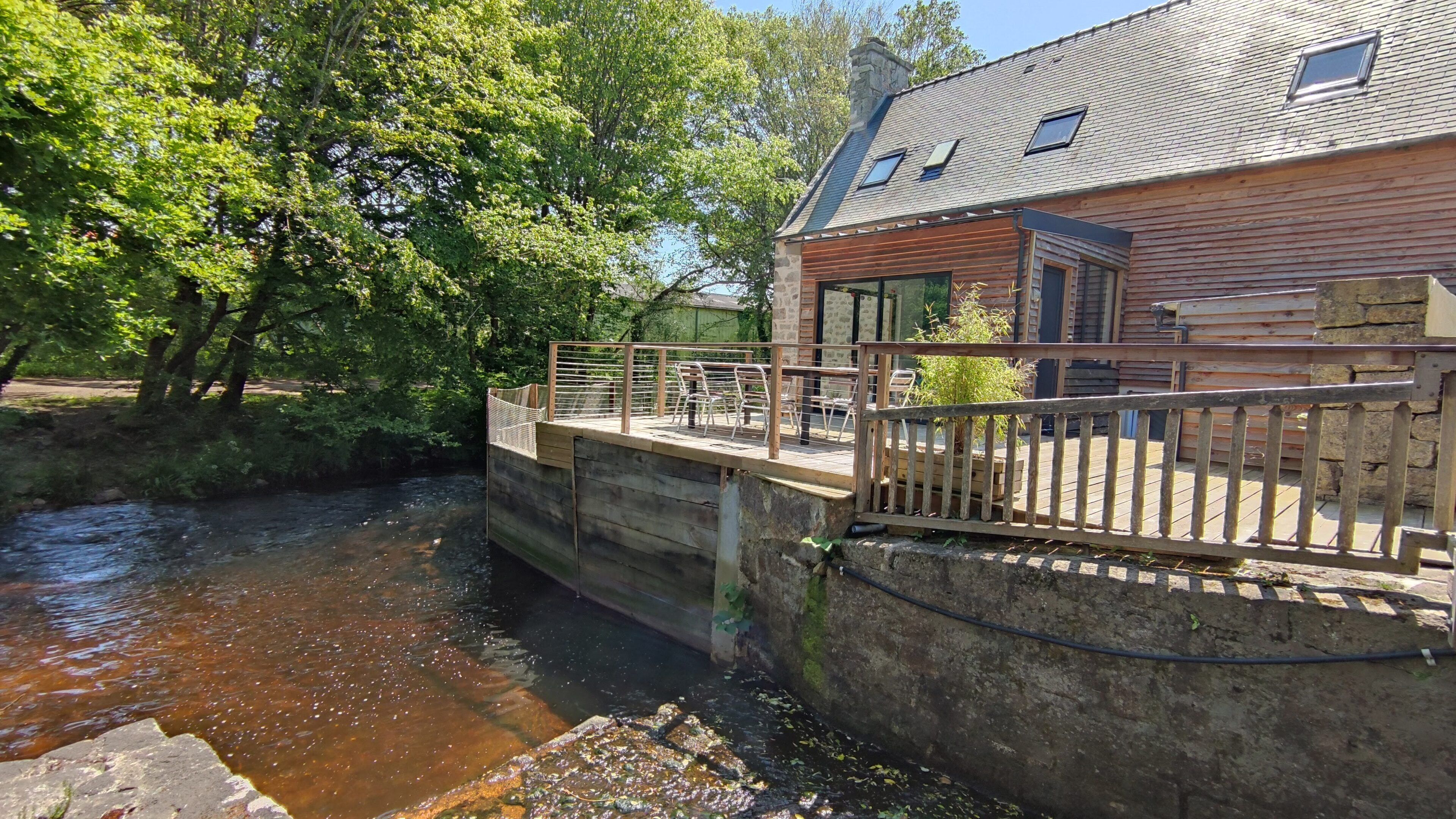 Eine Holzbrücke über einem Fluss, die zu einem Steinhaus mit schrägem Dach und Fenstern führt.