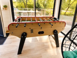 Una mesa de fútbolín en una habitación con ventanas grandes, una silla de ruedas y una planta en maceta.