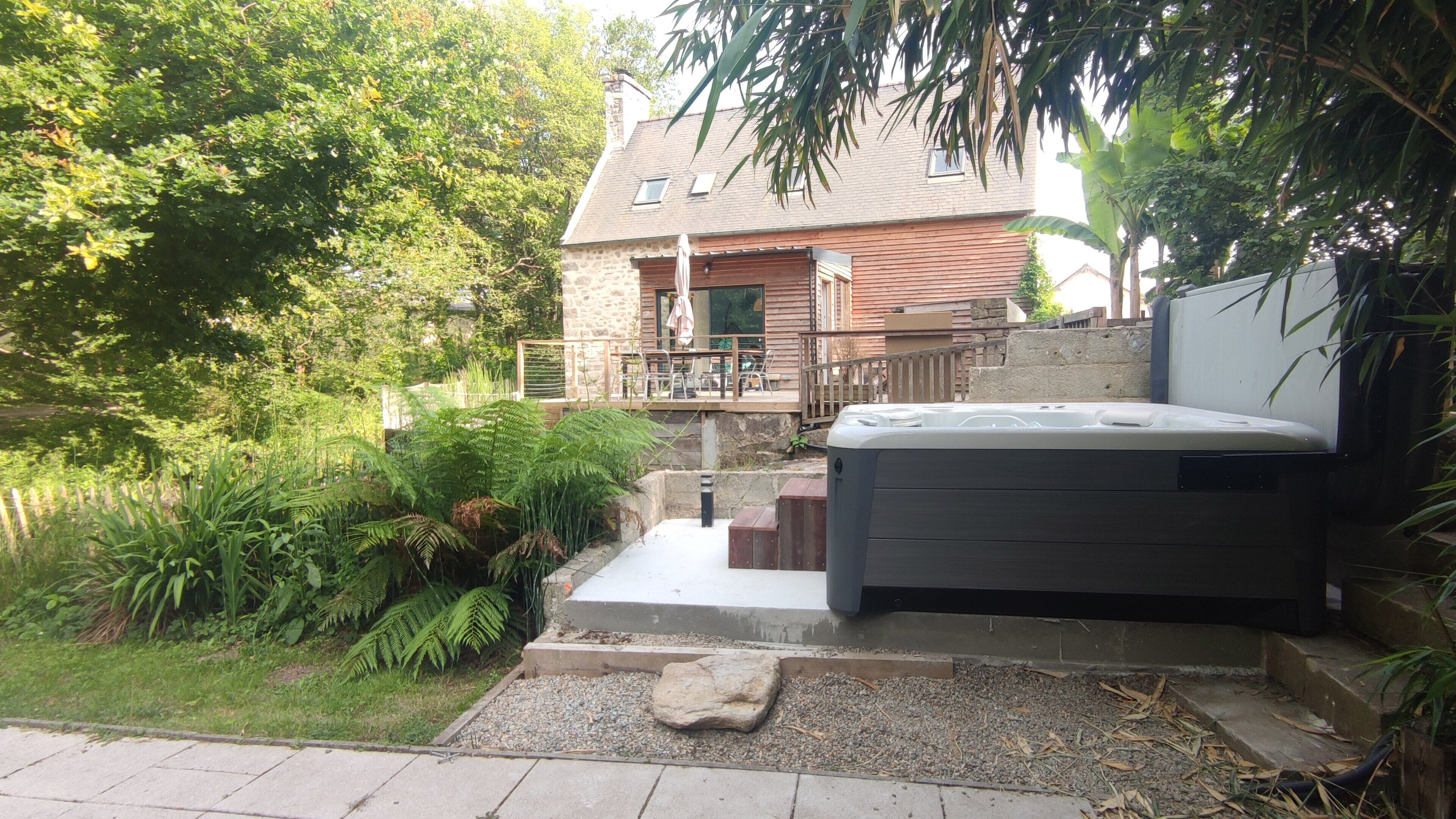 A patio area with a hot tub, a stone wall, and a wooden deck overlooking a garden.
