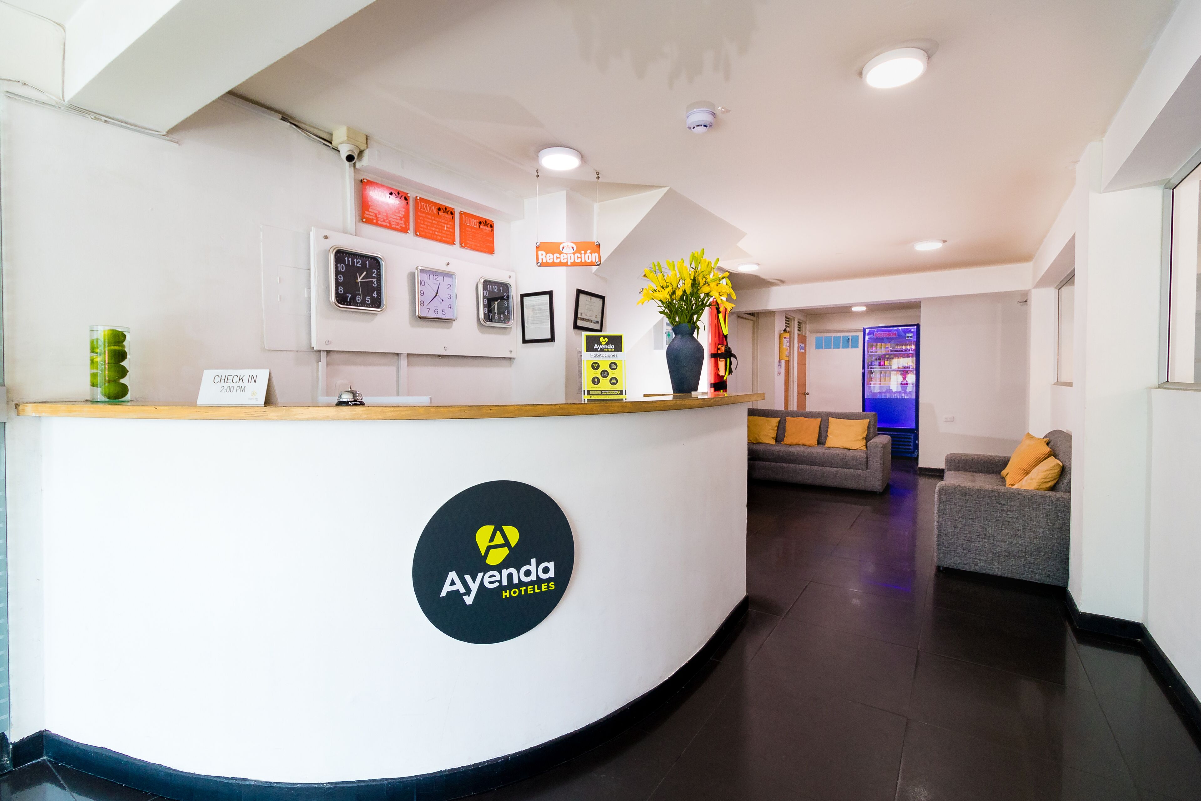 A hotel reception area with a reception desk, a clock, framed certificates, a sign indicating check-in time, and a vase with flowers.