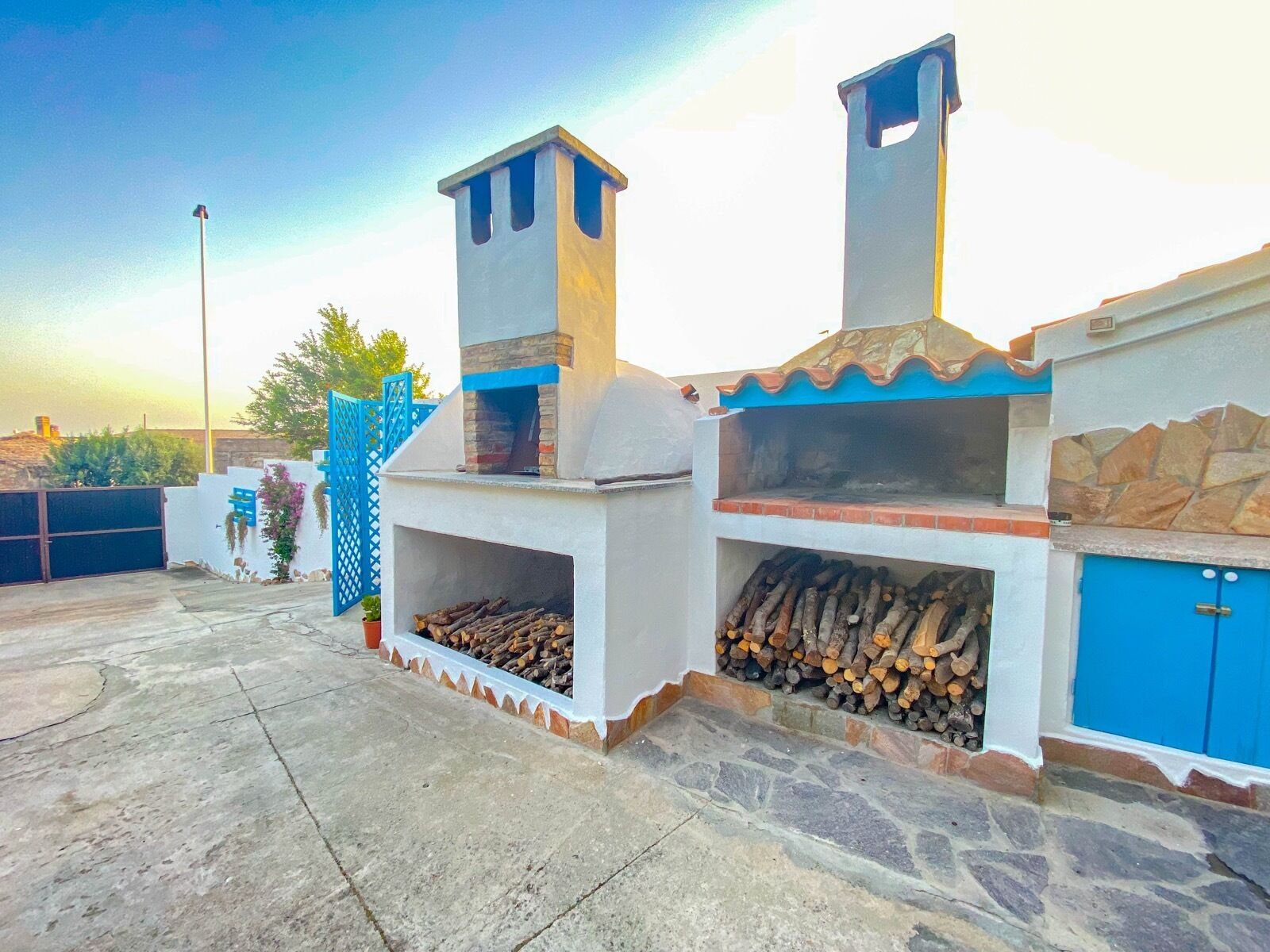 A stone and tile outdoor kitchen with two chimneys, a blue door, and a concrete patio.