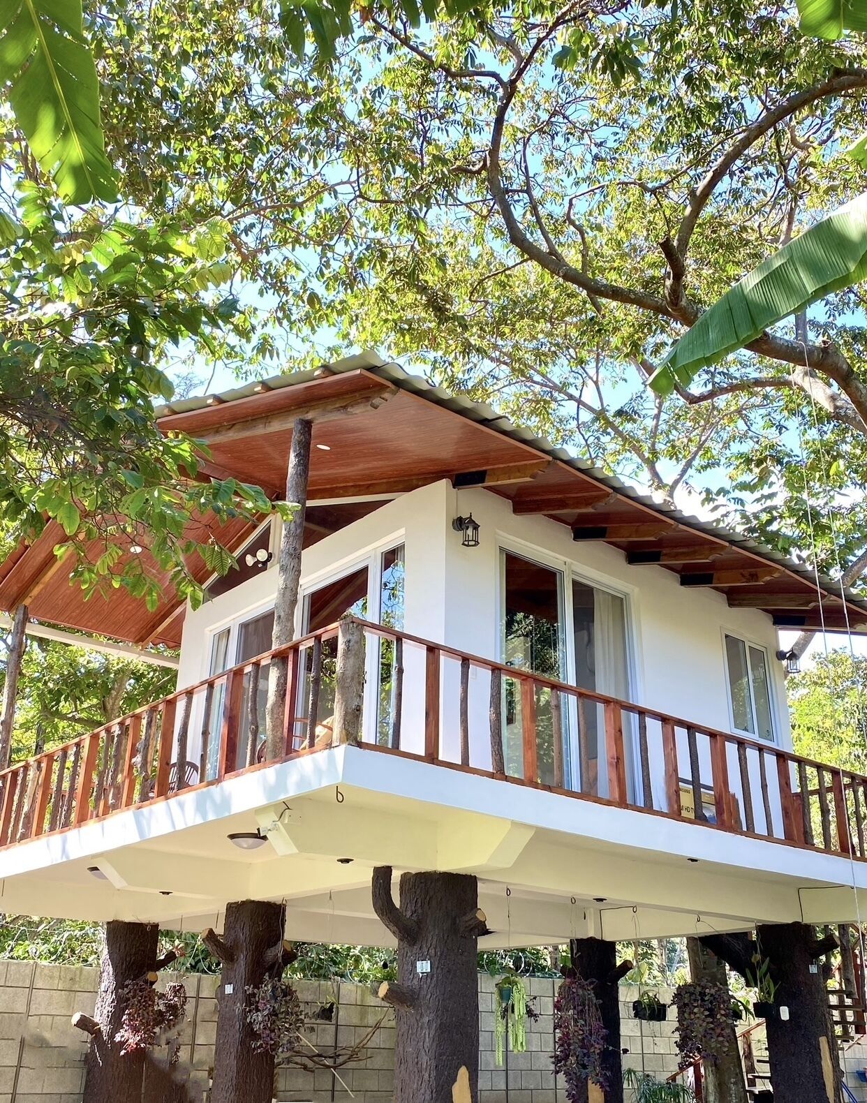 Une maison à deux étages dotée d’un balcon, soutenu par des troncs d’arbres, et entourée d’une végétation luxuriante.