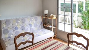 Une pièce avec un canapé adossé à un mur, une petite table en bois avec une lampe, et une fenêtre donnant sur du mobilier d’extérieur.