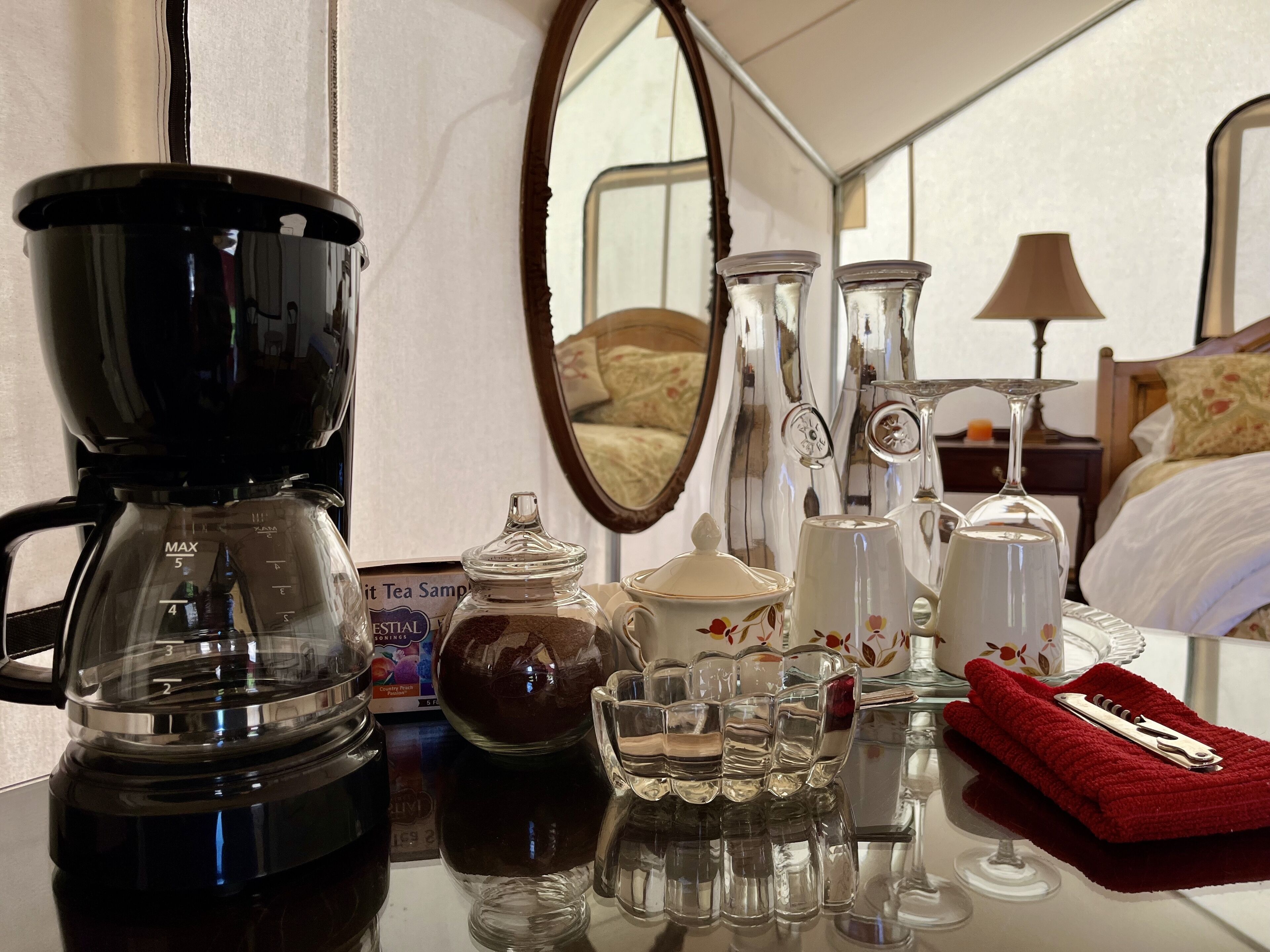 A coffee maker, a tea sampler, a glass jar, a set of glasses, and a red towel on a table in a hotel room.
