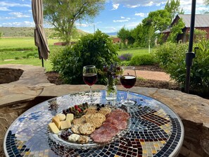 A table with a glass platter of cheese, crackers, and meats, two glasses of red wine, and a vase of flowers, set outdoors with a view of a rural landscape. - Beautiful Glamping Tent at Zenzen Gardens (Paonia)