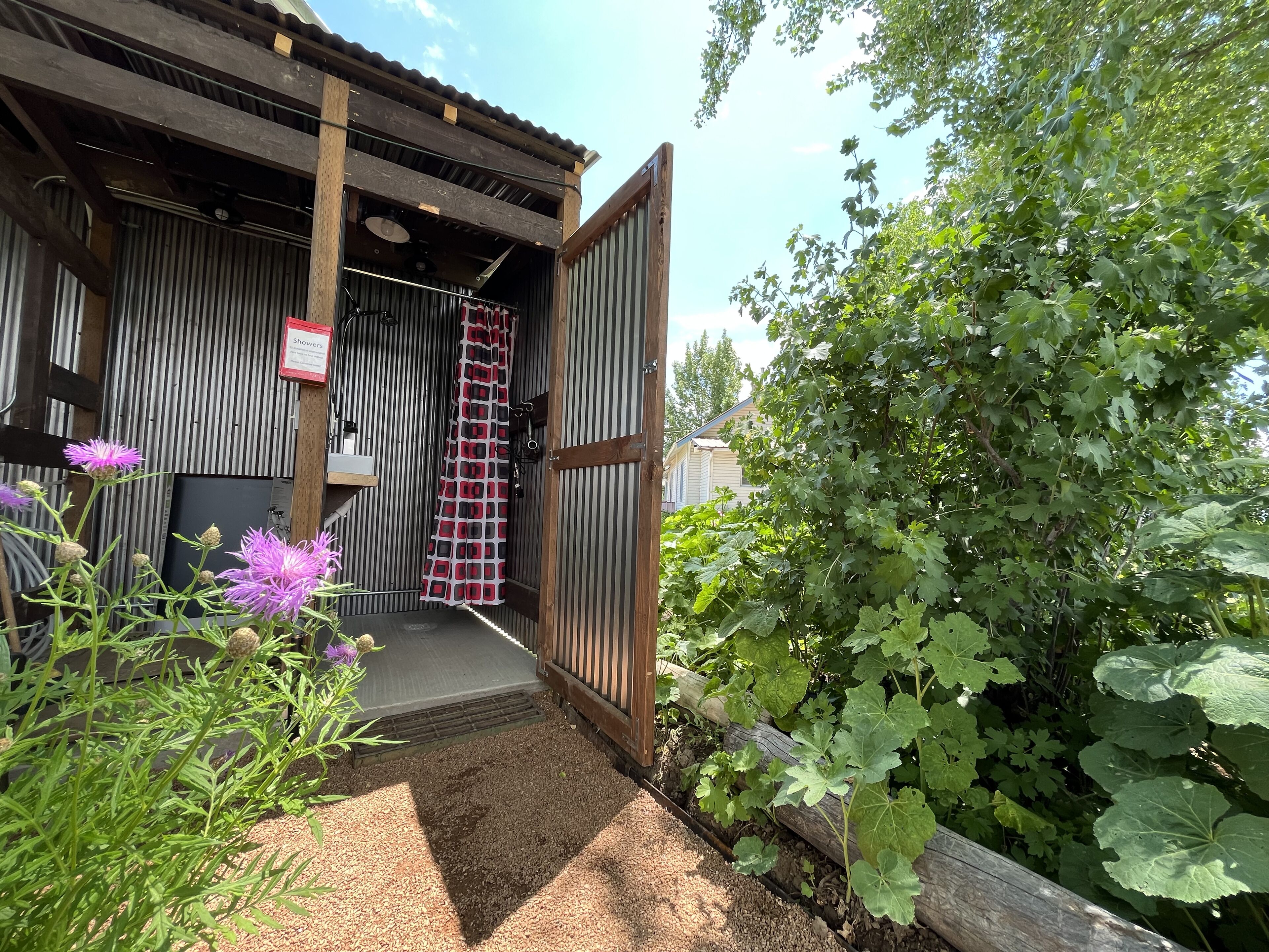 A rustic building with a wooden exterior, a metal door, and a garden area with green foliage and purple flowers.