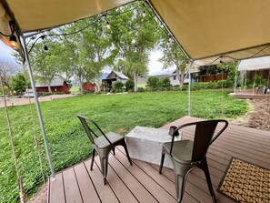 A covered patio with two chairs and a table, overlooking a well-maintained lawn and a rural backdrop with barns and houses. - Beautiful Glamping Tent at Zenzen Gardens (Paonia)
