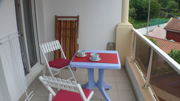 Un balcon avec une chaise blanche et une table, une fenêtre et une vue sur une maison et des arbres.