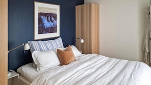 Een netjes opgemaakt bed met witte beddengoed en een enkel bruin kussen, een ingelijst kunstwerk erboven en een bedlamp op een houten nachtkastje.