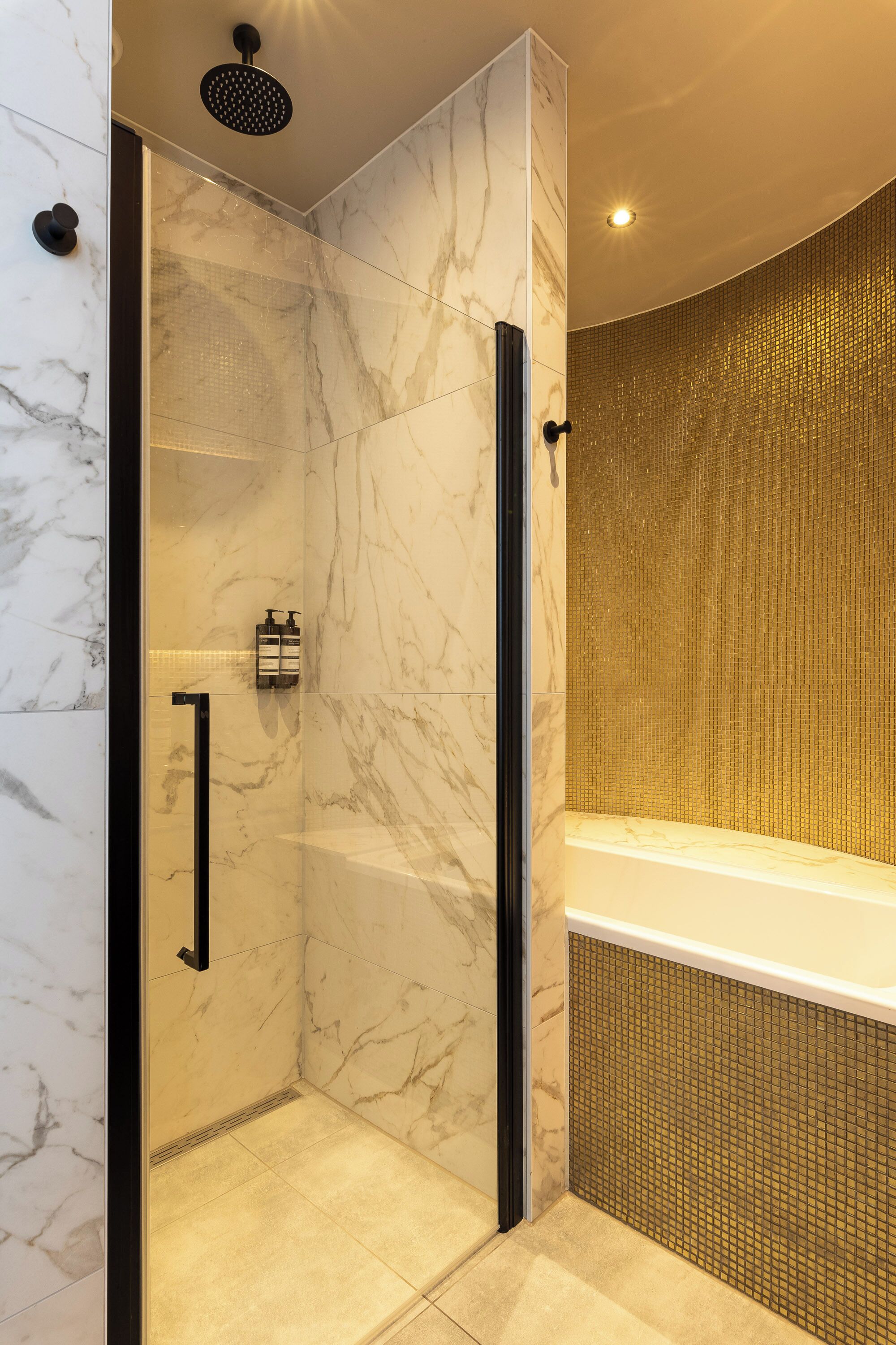 A modern bathroom with a glass-enclosed shower, marble walls, and a gold-tiled bathtub.