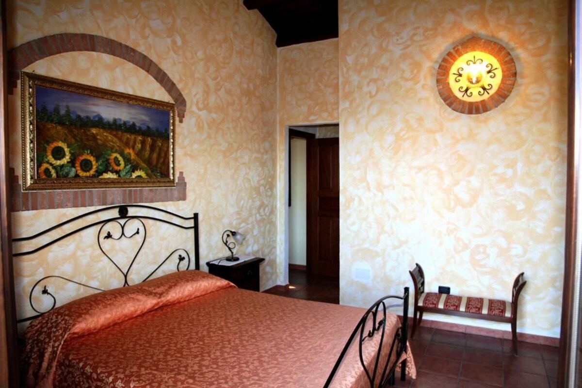 A bedroom with a bed, a nightstand, a bench, a painting, and a wall-mounted light fixture.