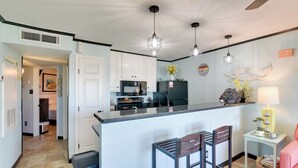 A modern kitchen with white cabinets, a central island with bar stools, pendant lighting, and a view into the adjacent living area. - Steps to Galveston Beach: Resort-Style Condo! (Galveston)