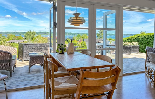 An open deck with a wooden table and chairs, a hanging light fixture, and a view of a green landscape through large windows.