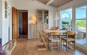 A dining area with a wooden table and chairs, a cabinet with glass doors, and a view through large windows.