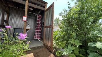 Un bâtiment rustique avec une façade en bois, une porte en métal et un jardin agrémenté de feuillage verdoyant et de fleurs violettes.