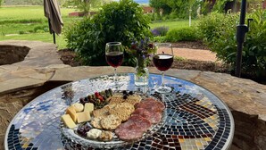 A table with a glass platter of cheese, crackers, and meats, two glasses of red wine, and a vase of flowers, set outdoors with a view of a rural landscape.