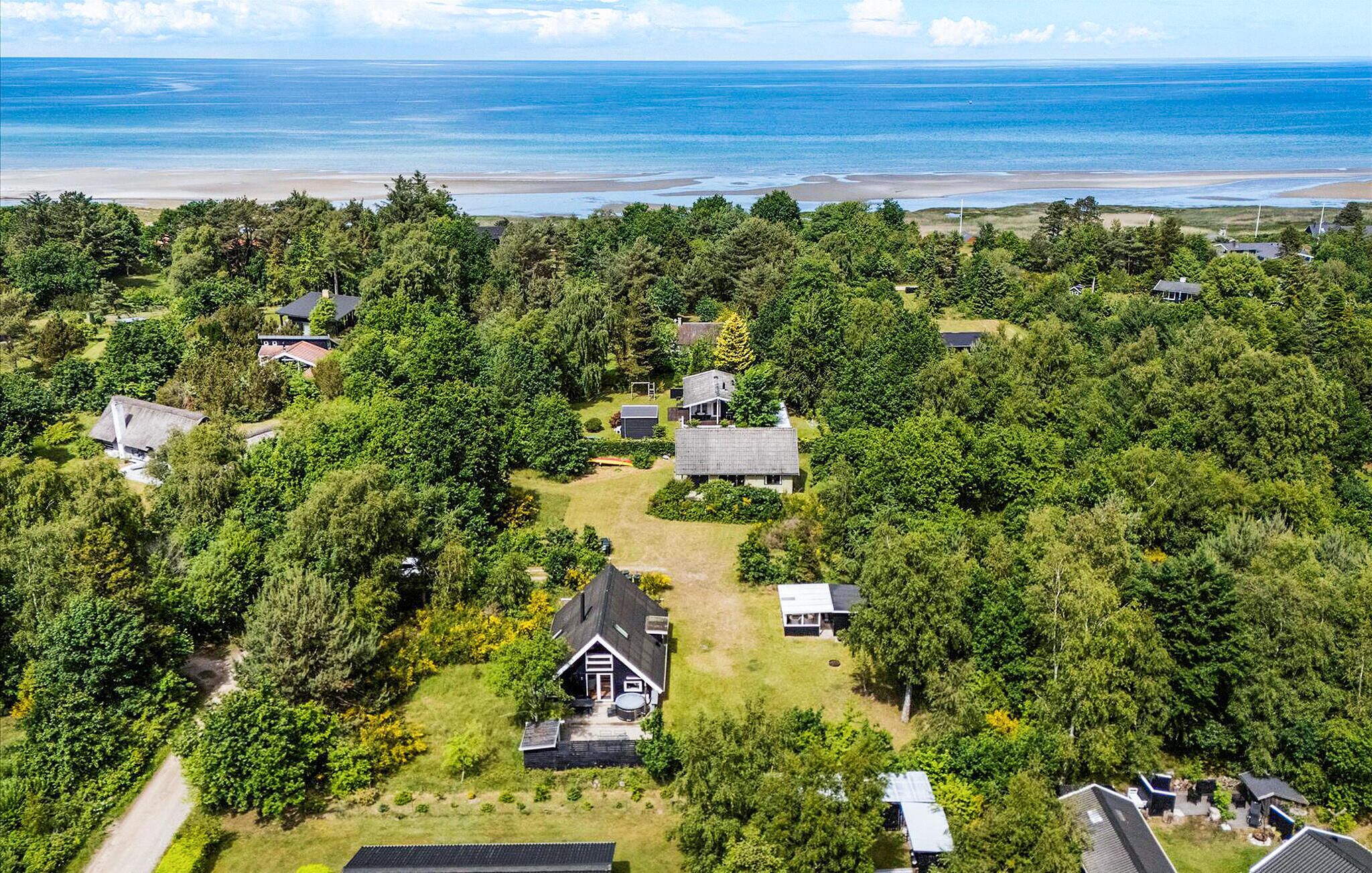 Une vue aérienne d’un quartier résidentiel, avec des maisons entourées d’une végétation luxuriante, une plage en arrière-plan et un ciel dégagé.