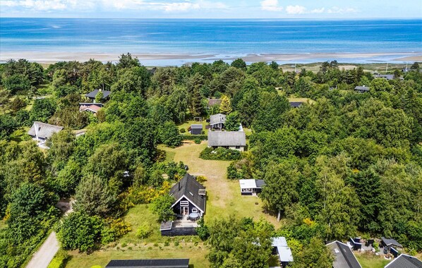 Luchtbeeld van een woongebied met huizen, omringd door weelderige begroeiing, een strand in de verte en een heldere lucht.