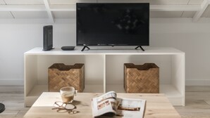 A modern living room with a flat-screen TV on a white stand, a coffee table with a magazine and glasses, and two woven baskets on a white console table. - Family holidays in Madeira - Casa da Lombada (Funchal)