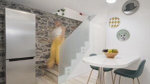 A modern kitchen with a stainless steel refrigerator, a stone accent wall, a white dining area with two green chairs, and a fruit bowl on the table. - Family holidays in Madeira - Casa da Lombada (Funchal)