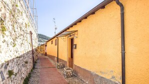 Une ruelle étroite, avec un bâtiment jaune d’un côté et un mur de pierre de l’autre. Des marches mènent à l’entrée du bâtiment, et on aperçoit une petite porte en bois.