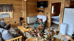 Interior acogedor de una cabaña con un sofá, una mesa con una cesta, un televisor montado en la pared y una cama al fondo.
