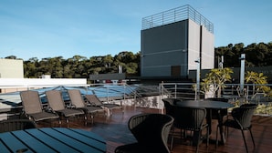 Um terraço no topo do prédio com espreguiçadeiras, mesas, uma piscina e o edifício ao fundo.