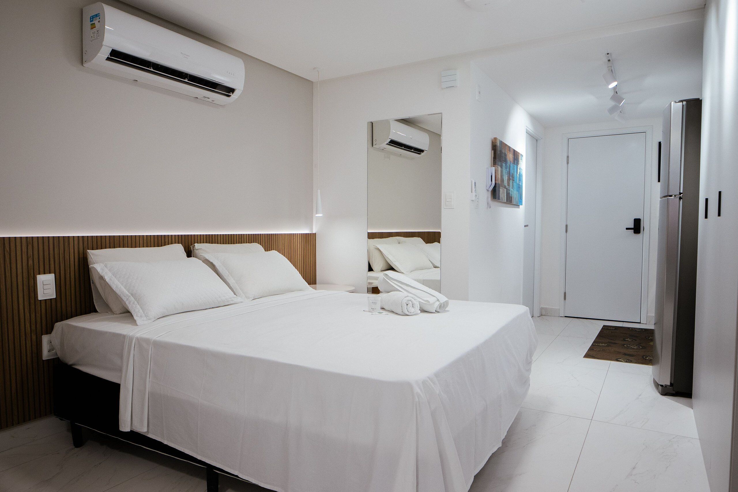 A hotel room with a large bed, a wardrobe, a mirror, an air conditioning unit, and a refrigerator.