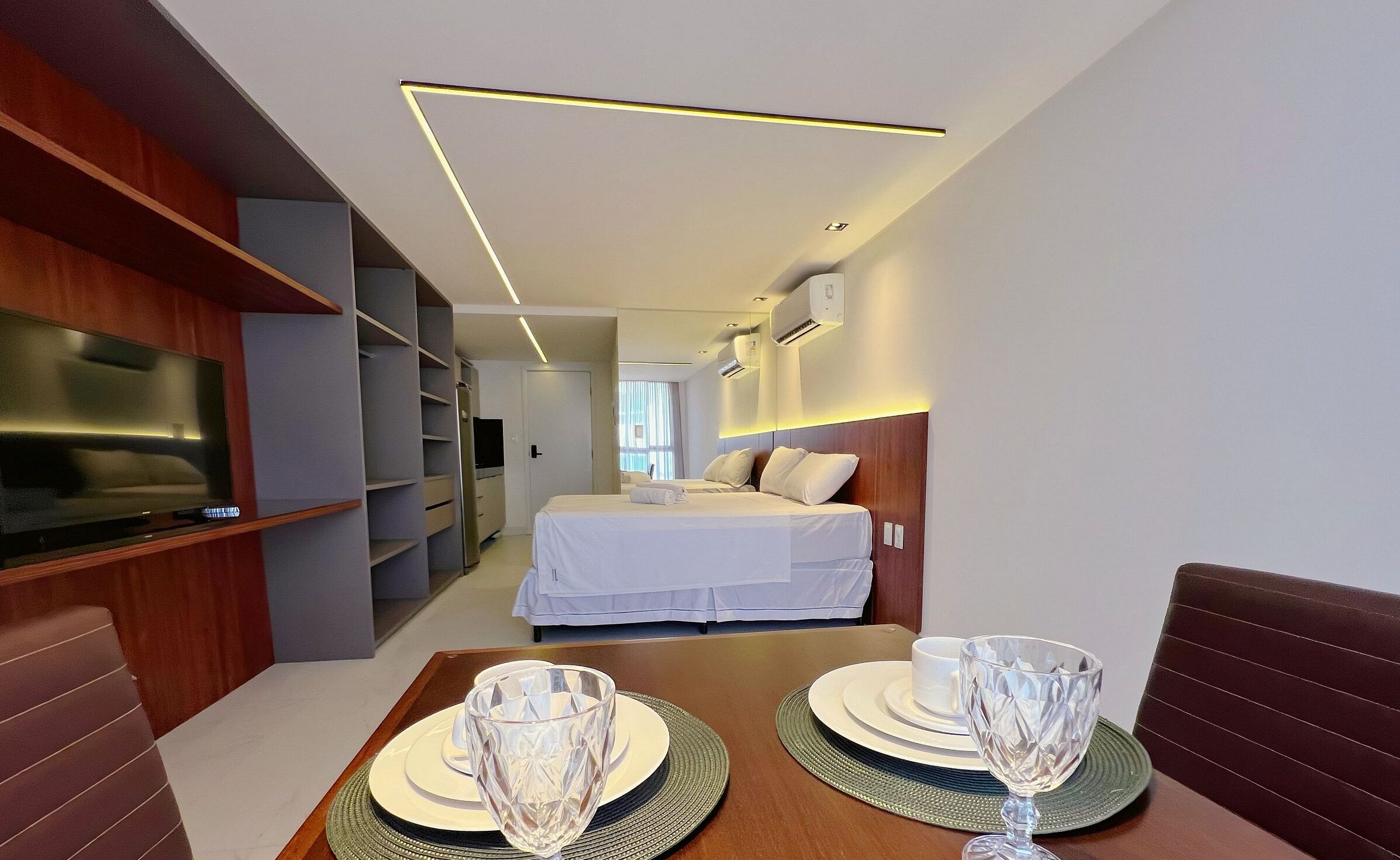 A modern hotel room with a TV, a bed, a dining table with glassware, and a walk-in closet.