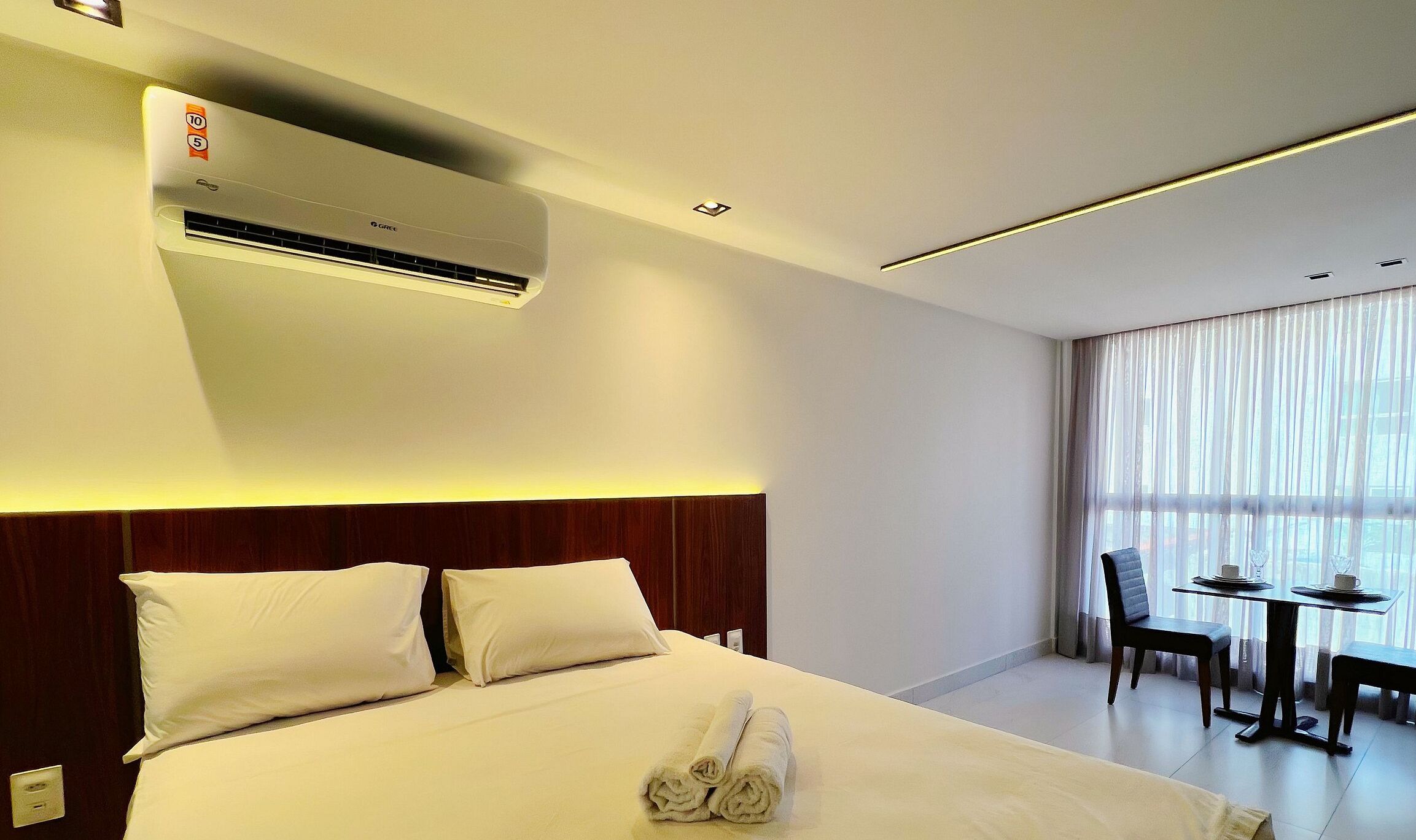 Quarto de hotel com cama, escrivaninha e cadeira. Há um aparelho de ar condicionado na parede.
