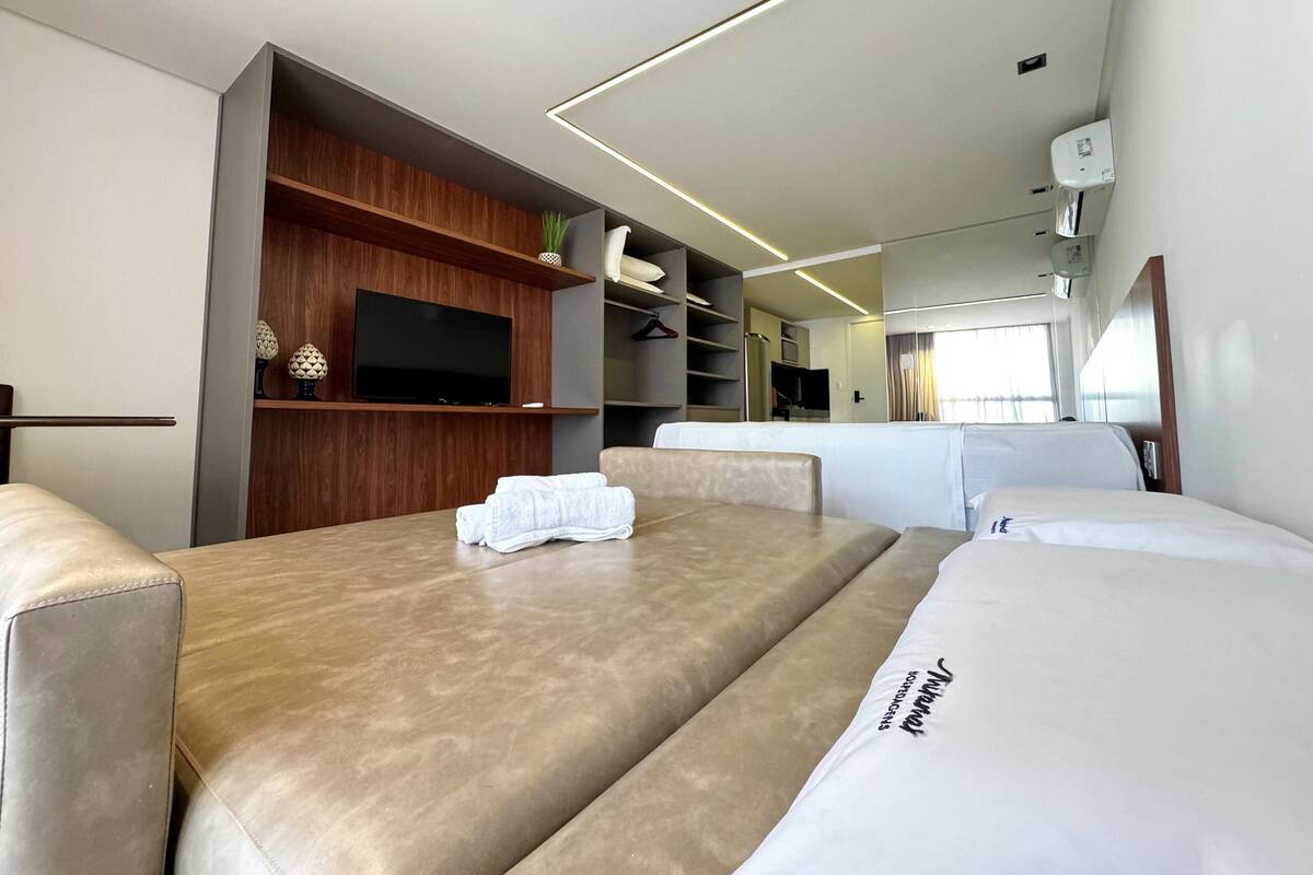 A modern hotel room with a large bed, a flat-screen TV, wooden shelving, and a bathroom with a hand dryer.