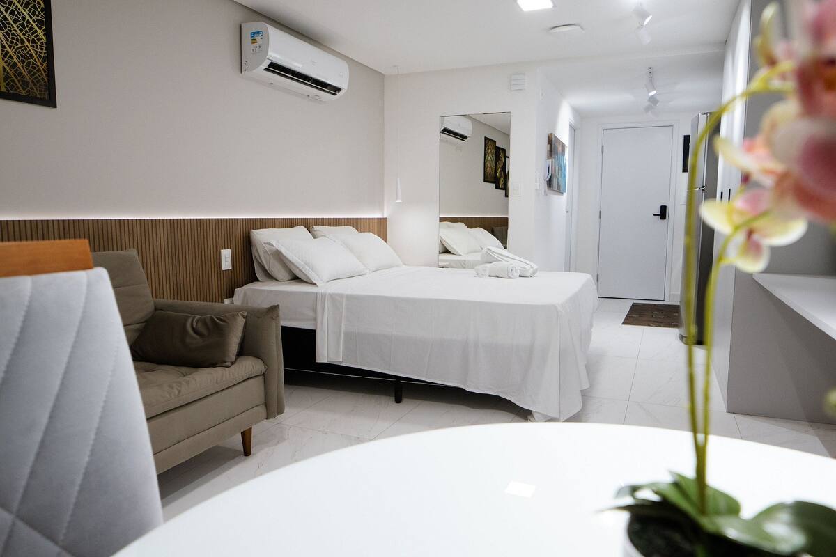 A modern hotel room with a bed, a sofa, a small table, and an air conditioning unit.