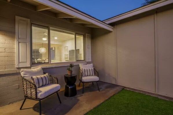 An outdoor seating area with two wicker chairs and a small table, a window with blinds, and a well-maintained lawn. - Biltmore 3 bedroom 2 bath super comfy property Great Location ! (Phoenix)