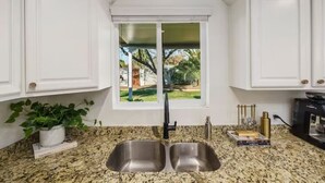 Cozinha com armários brancos, bancada de granito, pia dupla e janela com vista para o verde lá fora.