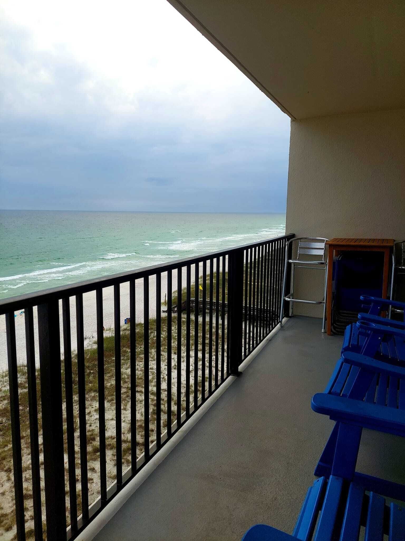 A balcony with a view of the beach and ocean. There are blue benches and a grill.