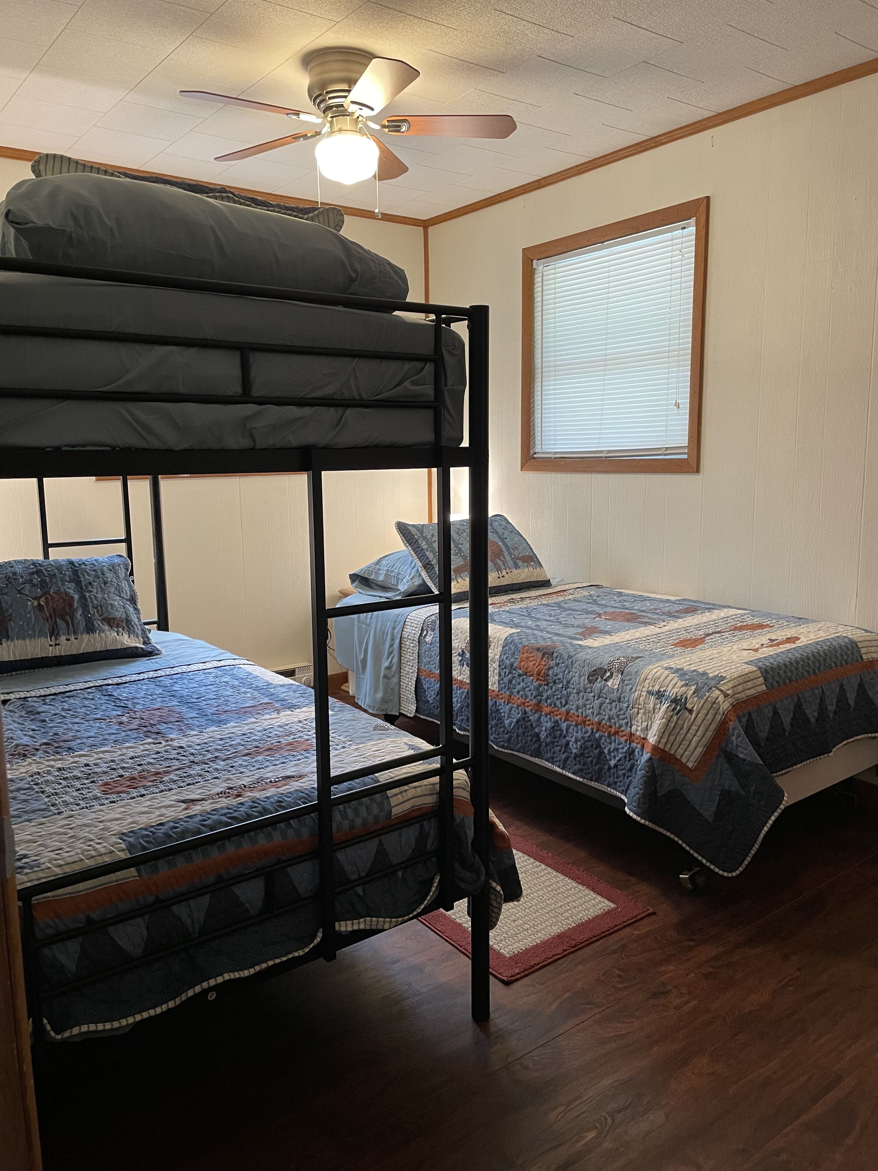 Une chambre avec des lits superposés, un ventilateur de plafond, une fenêtre avec des stores et un plancher en bois.