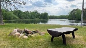 A picnic area with a stone table and fire pit surrounded by rocks, near a lake with trees in the background. - EAGLE LODGE - Large log cabin on the E. Fork of Chippewa River (Loretta)