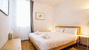 Un dormitorio ordenado con una cama de madera, ropa de cama blanca, una mesita de noche pequeña, un radiador y una ventana con cortinas.
