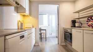 Una cocina moderna con armarios blancos, horno empotrado y cafetera. Se observa un comedor a través de una puerta abierta.