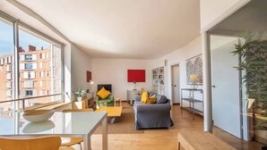 Un apartamento moderno con comedor, sala de estar con sofá y televisor, y un amplio ventanal con vistas a edificios.