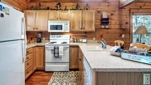 Una cocina tipo cabaña con armarios de madera, un refrigerador blanco, una estufa y un microondas. Hay elementos decorativos en los armarios, un lavamanos y una encimera con un cartel que dice "Cabin Guest Thank Life is Better at the Cabin".