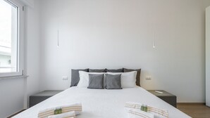 Un letto rifatto con cura, asciugamani bianchi e a righe, una testiera grigia e un comodino con un telecomando.