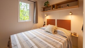 A bedroom with a bed, a window with curtains, a shelf with plants, and wall-mounted lights. - Le Palmier, mobile home not overlooked (Hyères)
