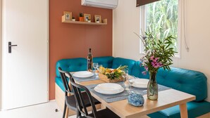 A dining area with a teal sofa, a table set for two, and a window with a view of greenery. - Le Palmier, mobile home not overlooked (Hyères)